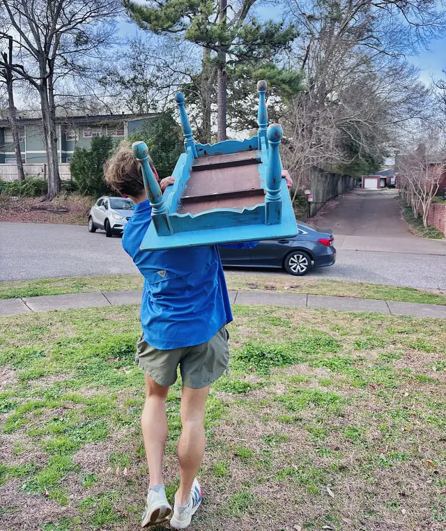 Mover carrying a blue chair overhead across a yard