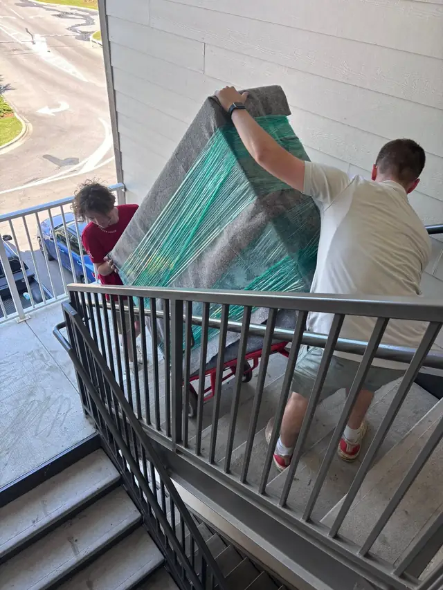 Two movers carrying wrapped furniture along an apartment balcony